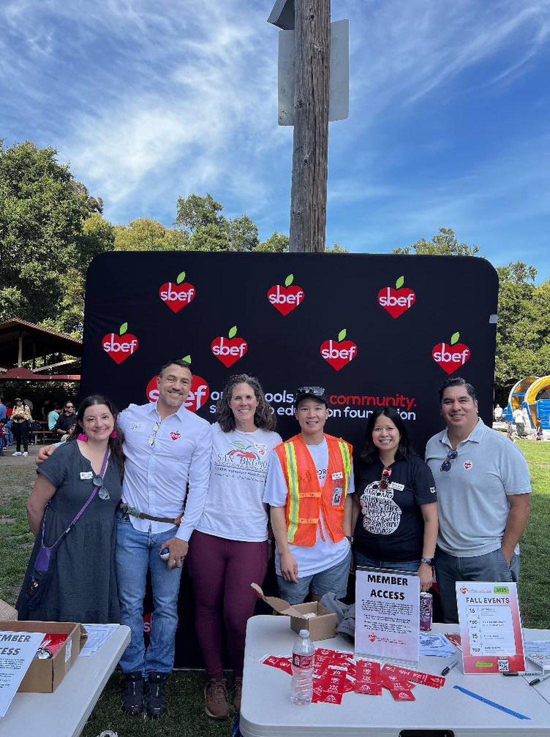 Six SBEF board members standing at a branded check-in table at the 2025 Back to School BBQ, with member-access signage, a fall events board, name badges and a volunteer in a hi-vis vest