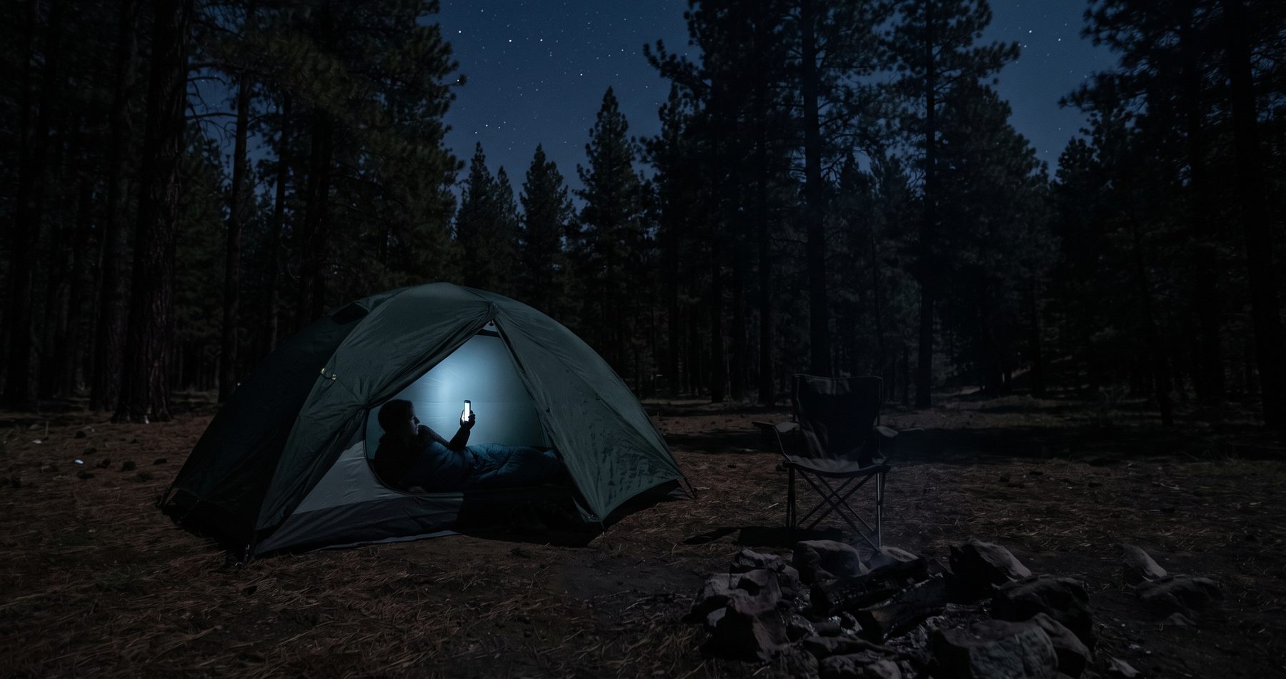 A tent at a dark campsite at night, lit from inside by the glow of a phone screen held by someone lying in a sleeping bag