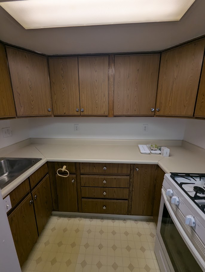 Vacant rental kitchen with dated brown laminate cabinets, dropped fluorescent ceiling, and diamond vinyl floor — before renovation